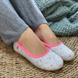 A pair of gray slippers with pink piping and heart shapes on the uppers, worn by a person sitting with their legs visible, wearing gray pants and a green sweater. Cosy comfortable Spanish slippers/house shoe.